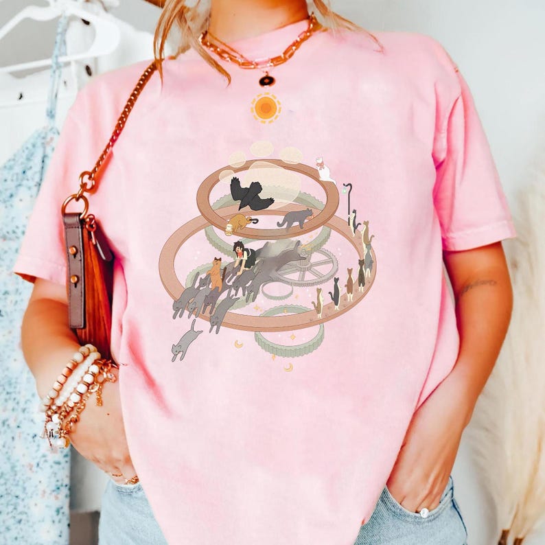 May include: A light pink t-shirt featuring a whimsical illustration of cats. The design includes multiple cats, a sun, and circular elements. The shirt is a comfortable, casual style.