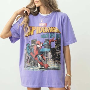 Puede incluir: Una camiseta lavanda con un gr&aacute;fico de Spider-Man. El dise&ntilde;o incluye el logotipo de Marvel, las palabras "Spider-Man" en amarillo y "Brand New Day" en blanco. El gr&aacute;fico representa a Spider-Man en una pose din&aacute;mica contra un horizonte urbano.