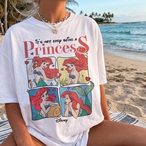 May include: A white short-sleeve t-shirt featuring a graphic print of the character Ariel from The Little Mermaid in four distinct panels. The text reads It's not easy being a Princess and includes the Disney logo at the bottom of the graphic.