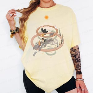 May include: Light yellow t-shirt featuring a whimsical illustration of cats within a circular design. The graphic includes a sun, gears, and various cat silhouettes. The shirt has short sleeves and a relaxed fit.