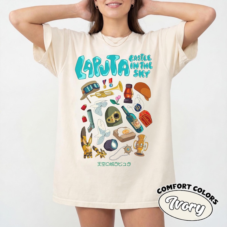 May include: Ivory-colored t-shirt featuring a colorful graphic design with the text "LAPUTA" and "CASTLE IN THE SKY". The design includes various illustrations of objects and characters. The shirt also has the text "COMFORT COLORS" and "Ivory".