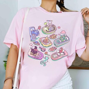 K&ouml;nnte beinhalten: Hellrosa T-Shirt mit einem farbenfrohen Grafikdesign von animierten Charakteren in Mobiltelefonen. Das Design zeigt Charaktere aus beliebten Zeichentrickfilmen und -serien. Das T-Shirt besteht aus weichem Material.
