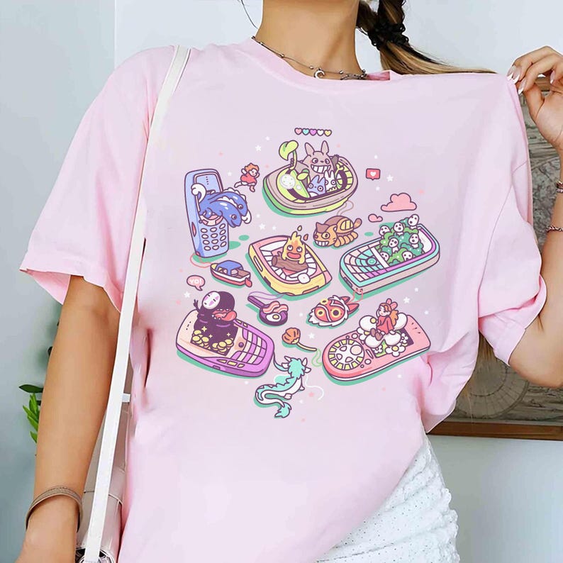 K&ouml;nnte beinhalten: Hellrosa T-Shirt mit einem farbenfrohen Grafikdesign von animierten Charakteren in Mobiltelefonen. Das Design zeigt Charaktere aus beliebten Zeichentrickfilmen und -serien. Das T-Shirt besteht aus weichem Material.