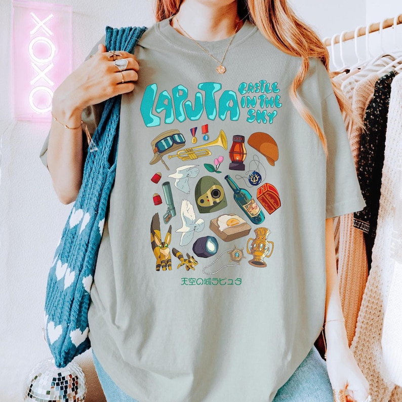 May include: A light green t-shirt featuring a collection of cartoon illustrations from "Castle in the Sky." The shirt has the words "LAPUTA" and "CASTLE IN THE SKY" in blue. Additional Japanese text is also present.