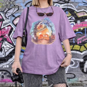 Puede incluir: Camiseta morada con un dibujo animado de un personaje cocinando en una sart&eacute;n. La camiseta tiene mangas cortas y un corte holgado. El texto "ORCHID COMFORT COLORS" est&aacute; impreso en la parte superior izquierda.