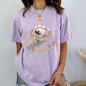 May include: A light purple t-shirt featuring a whimsical cat-themed graphic. The design includes a sun, paw prints, and a circular arrangement of cats. The shirt is made of a soft material and has short sleeves.