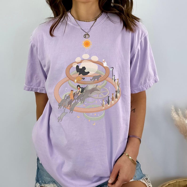 May include: A light purple t-shirt featuring a whimsical cat-themed graphic. The design includes a sun, paw prints, and a circular arrangement of cats. The shirt is made of a soft material and has short sleeves.