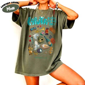 May include: Moss green t-shirt featuring a colorful graphic design with the text "Laputa Castle in the Sky." The design includes various illustrations such as a trumpet, lantern, and a bottle. The shirt is a comfort colors style.
