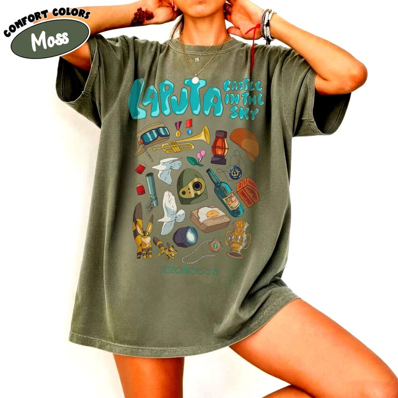May include: Moss green t-shirt featuring a colorful graphic design with the text "Laputa Castle in the Sky." The design includes various illustrations such as a trumpet, lantern, and a bottle. The shirt is a comfort colors style.