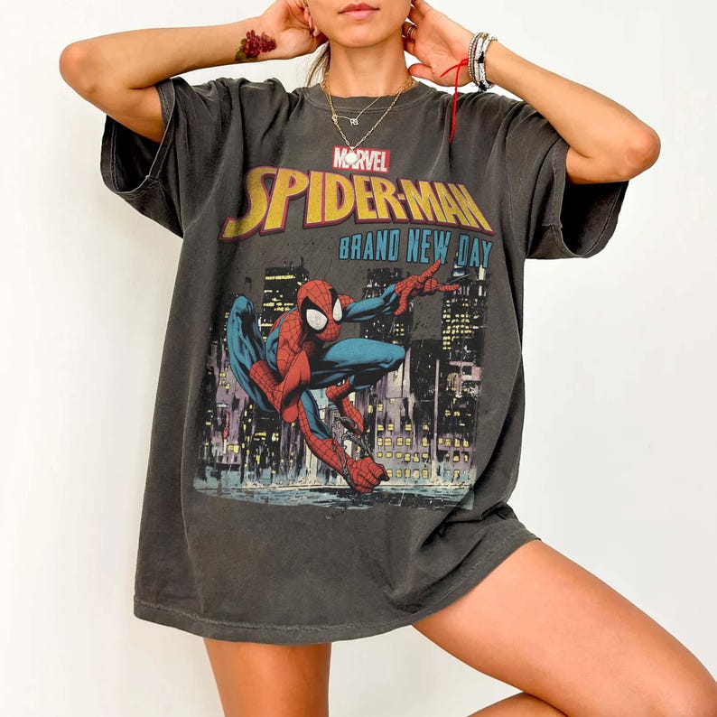 Puede incluir: Camiseta gris oscuro con un gr&aacute;fico de Spider-Man frente a un paisaje urbano. El logotipo de Marvel y las palabras "SPIDER-MAN BRAND NEW DAY" son amarillas y azules. La camiseta es de talla grande.