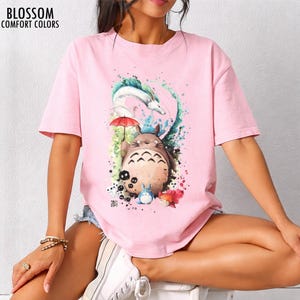 以下が含まれることがあります： ピンク色のTシャツ。大きな毛むくじゃらの生き物、小さな青い生き物、そしてドラゴンが描かれたカラフルなグラフィック。Tシャツの左上には「BLOSSOM COMFORT COLORS」の文字。