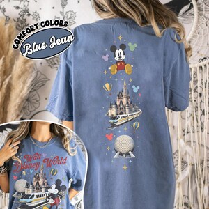 Pu&ograve; includere: Una t-shirt color blu jeans con un design grafico verticale di Topolino, un castello, una monorotaia e una sfera geodetica. Il testo COMFORT COLORS e Blue Jean appare in un badge circolare. Il design include piccole stelle e icone.
