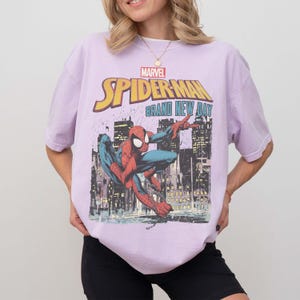 Puede incluir: Camiseta lila claro con un dise&ntilde;o de Spider-Man de Marvel. El gr&aacute;fico muestra a Spider-Man con el texto "Spider-Man Brand New Day" sobre un fondo de ciudad. La camiseta es de color lila claro.