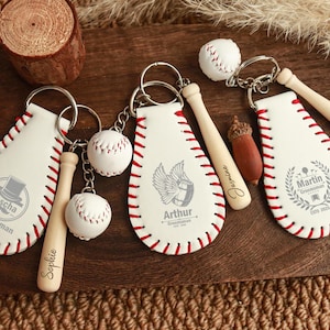 Könnte beinhalten: Drei weiße Schlüsselanhänger aus Leder in Baseballform mit roten Nähten. Jeder verfügt über einen Metallring, einen kleinen Baseballschläger aus Holz und einen Miniatur-Baseball. Die Gravuren zeigen Namen wie Jascha, Arthur, Martin, Sophie, Jasmine und Kristine.