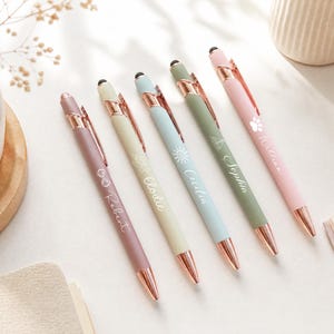 Könnte beinhalten: Fünf einziehbare Kugelschreiber mit matten Gehäusen in Mauve, Creme, Hellblau, Salbeigrün und Rosa. Jeder Stift verfügt über roségoldene Akzente, einen Clip und einen in weißer Schreibschrift eingravierten Namen neben einem kleinen Symbol.