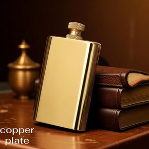 Puede incluir: Un frasco pulido de color dorado con una tapa texturizada. El frasco es rectangular y tiene una superficie brillante. Las palabras "copper plate" est&aacute;n en la parte inferior de la imagen. Una pila de libros encuadernados en cuero marr&oacute;n est&aacute; en el fondo.