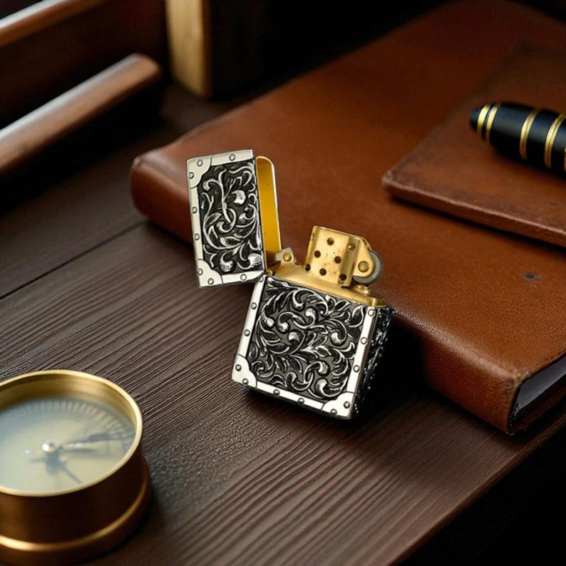 Peut inclure: Briquet ouvrag&eacute; avec une finition argent&eacute;e et dor&eacute;e. Le briquet pr&eacute;sente des volutes florales complexes sur les panneaux argent&eacute;s. Le dessus est ouvert, r&eacute;v&eacute;lant l'int&eacute;rieur dor&eacute;. Une boussole est visible au premier plan.