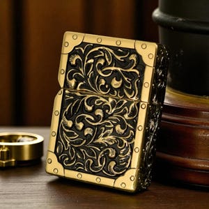 Peut inclure: Un briquet en m&eacute;tal dor&eacute; avec un motif floral noir. Le briquet rectangulaire pr&eacute;sente des coins arrondis et de petits rivets circulaires. Le design d&eacute;taill&eacute; lui donne un aspect vintage. Un accessoire classique.