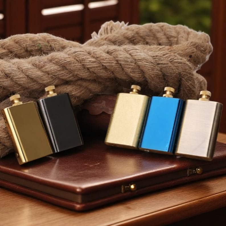 Puede incluir: Una colecci&oacute;n de petacas rectangulares en varios colores, incluyendo dorado, negro, azul y plateado. Cada petaca tiene una peque&ntilde;a tapa dorada. Las petacas est&aacute;n dispuestas sobre una superficie de cuero marr&oacute;n, con una gruesa cuerda en el fondo.