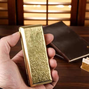 May include: A gold-coloured metal money clip with an embossed floral design. The rectangular clip is held in a hand, with a dark brown notebook in the background. The clip's design suggests a vintage or classic aesthetic, with a polished finish.