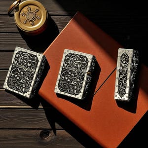 Peut inclure: Trois briquets argent&eacute;s avec des motifs floraux noirs orn&eacute;s. Les briquets sont rectangulaires avec des coins arrondis et un bord argent&eacute;. Ils sont dispos&eacute;s sur une surface en cuir marron, avec une boussole en laiton en arri&egrave;re-plan.
