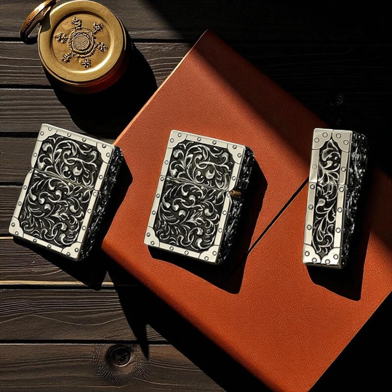 Peut inclure: Trois briquets argent&eacute;s avec des motifs floraux noirs orn&eacute;s. Les briquets sont rectangulaires avec des coins arrondis et un bord argent&eacute;. Ils sont dispos&eacute;s sur une surface en cuir marron, avec une boussole en laiton en arri&egrave;re-plan.