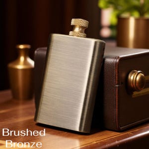 Puede incluir: Petaca de bronce cepillado con acabado texturizado y tap&oacute;n dorado. La petaca es rectangular y se apoya en una caja de cuero marr&oacute;n. Las palabras "Brushed Bronze" est&aacute;n impresas en la parte inferior de la imagen.