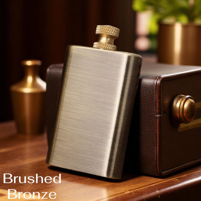 Puede incluir: Petaca de bronce cepillado con acabado texturizado y tap&oacute;n dorado. La petaca es rectangular y se apoya en una caja de cuero marr&oacute;n. Las palabras "Brushed Bronze" est&aacute;n impresas en la parte inferior de la imagen.