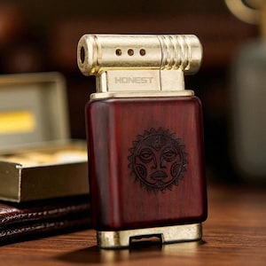May include: A rectangular lighter with a gold-coloured top and base, and a dark reddish-brown wooden body. The word "HONEST" is printed on the top. A sun-like face is carved into the wood. The lighter is on a wooden surface.