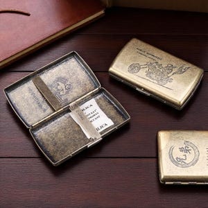 Puede incluir: Cajetilla de cigarrillos de latón antiguo con diseño de motocicleta Harley-Davidson. La caja está abierta, revelando un compartimento y un pequeño paquete. La caja es rectangular con esquinas redondeadas y tapa con bisagras. Sobre una superficie de madera oscura.