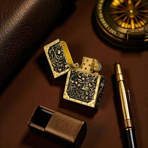 Peut inclure: Un briquet de style ancien, ouvert, avec des d&eacute;tails floraux noirs et dor&eacute;s complexes. Le briquet est pos&eacute; sur une surface marron, &agrave; c&ocirc;t&eacute; d'un &eacute;tui assorti, d'un stylo dor&eacute; et d'une boussole. L'int&eacute;rieur du briquet est dor&eacute;.