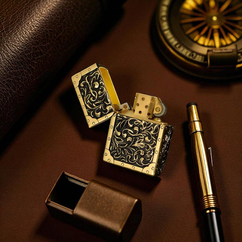 Peut inclure: Un briquet de style ancien, ouvert, avec des d&eacute;tails floraux noirs et dor&eacute;s complexes. Le briquet est pos&eacute; sur une surface marron, &agrave; c&ocirc;t&eacute; d'un &eacute;tui assorti, d'un stylo dor&eacute; et d'une boussole. L'int&eacute;rieur du briquet est dor&eacute;.