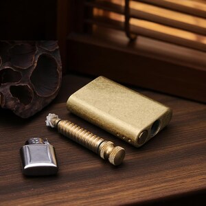 Puede incluir: Un conjunto de accesorios para fumar, que incluye una pipa rectangular de lat&oacute;n, una herramienta de lat&oacute;n y un encendedor plateado. Los art&iacute;culos est&aacute;n dispuestos sobre una superficie de madera oscura, con un fondo borroso.