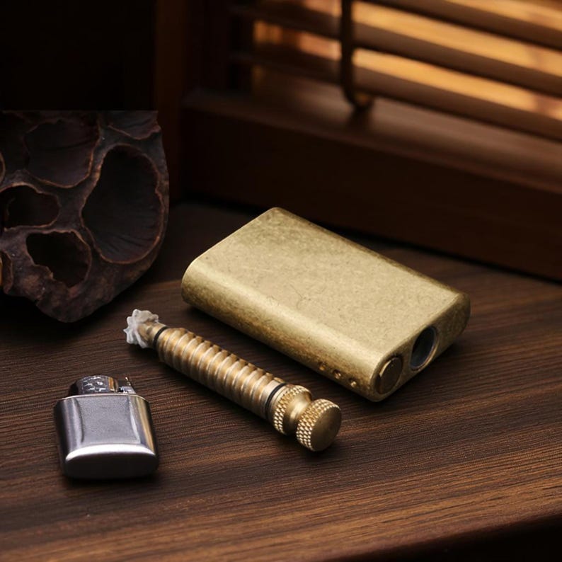 Puede incluir: Un conjunto de accesorios para fumar, que incluye una pipa rectangular de lat&oacute;n, una herramienta de lat&oacute;n y un encendedor plateado. Los art&iacute;culos est&aacute;n dispuestos sobre una superficie de madera oscura, con un fondo borroso.