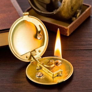 Puede incluir: Un encendedor de bolsillo redondo de color dorado, con la tapa abierta, que revela el mecanismo del encendedor. Una llama está encendida, con el encendedor apoyado sobre una superficie de madera. El encendedor tiene una tapa con bisagras y un pequeño indicador de combustible.