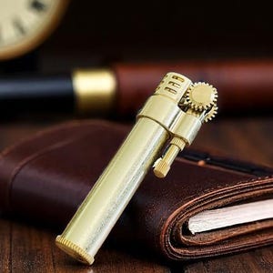 May include: A gold-coloured, vintage-style lighter with a gear mechanism on top. The lighter is cylindrical with a textured surface and is resting on a brown leather notebook. The text "Original Copper" is visible.
