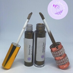 Könnte beinhalten: Zwei Flaschen bronzierter Lipgloss mit Applikatoren. Ein Applikator hat eine goldene Kappe, der andere eine roségoldene Kappe mit weißen Perlenakzenten. Die Flaschen haben weiße Etiketten mit dem Text "Bronzed Treat" und einem Nettogewicht von 10 ml.