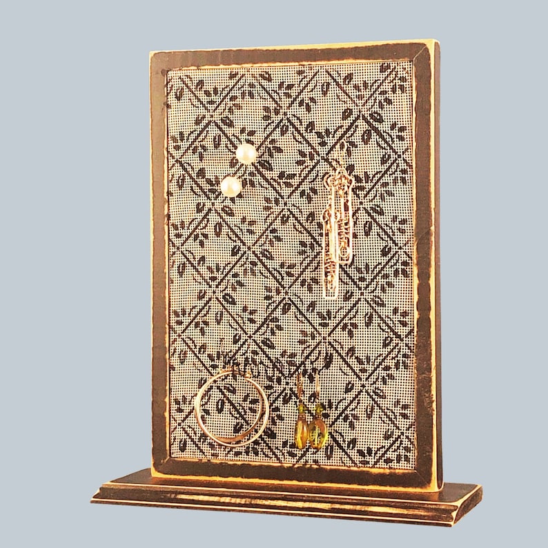 Puede incluir: Un expositor de joyas de madera con un fondo estampado en marr&oacute;n y negro. El expositor tiene un peque&ntilde;o estante en la parte inferior y cuenta con ganchos y un peque&ntilde;o estante para exhibir pendientes, collares y pulseras.