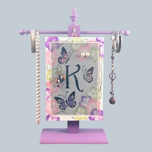 May include: Purple jewelry stand with a butterfly and floral design. The stand has a mesh panel for hanging earrings and a rod for bracelets. The stand also has a base with the letter "K" painted on it.