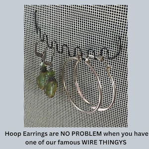 May include: A black metal wire earring organizer rack featuring a wavy design for hanging jewelry. Two pairs of earrings are displayed on the rack: one pair of green teardrop-shaped earrings and one pair of large silver hoop earrings. Text reads: Hoop Earrings are NO PROBLEM when you have one of our famous WIRE THINGYS.