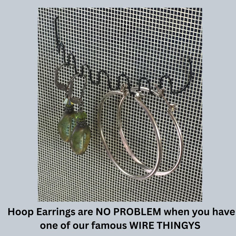 May include: A black metal wire earring organizer rack featuring a wavy design for hanging jewelry. Two pairs of earrings are displayed on the rack: one pair of green teardrop-shaped earrings and one pair of large silver hoop earrings. Text reads: Hoop Earrings are NO PROBLEM when you have one of our famous WIRE THINGYS.