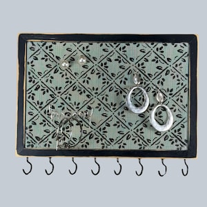 May include: A rectangular jewelry organizer with a black frame and a light green mesh backing featuring a black diamond leaf pattern. The board holds two pearl stud earrings, two silver hoop earrings, and two silver oval drop earrings.