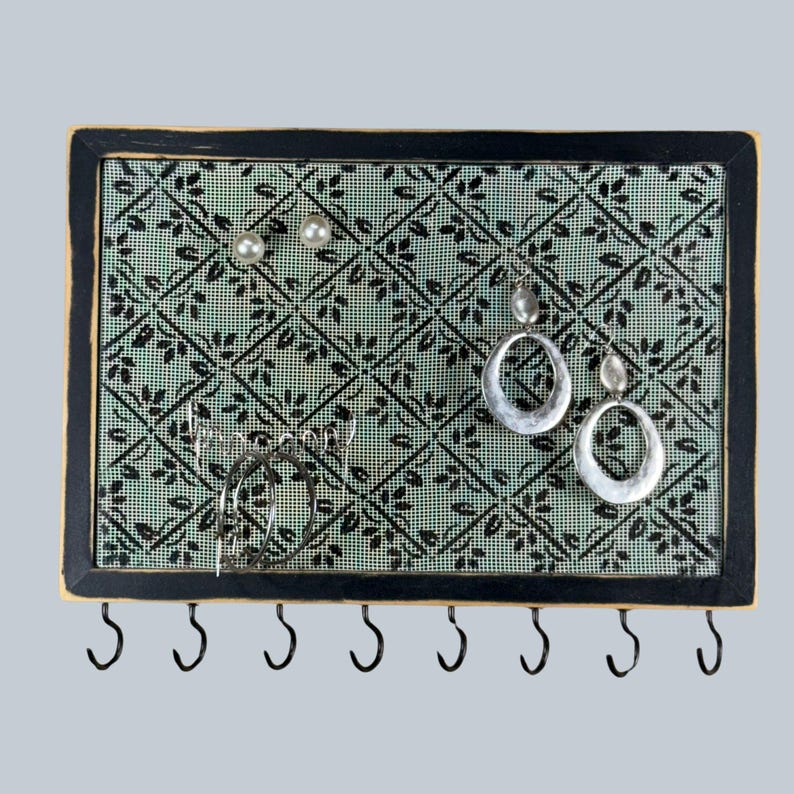 May include: A rectangular jewelry organizer with a black frame and a light green mesh backing featuring a black diamond leaf pattern. The board holds two pearl stud earrings, two silver hoop earrings, and two silver oval drop earrings.