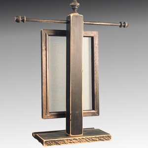 Puede incluir: Un expositor de joyas de madera negra con un marco rectangular y una varilla de metal. El expositor tiene un acabado desgastado y un peque&ntilde;o remate en la parte superior.
