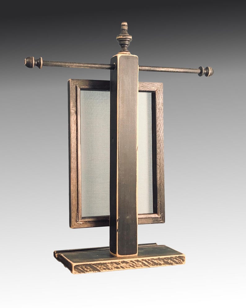 Puede incluir: Un expositor de joyas de madera negra con un marco rectangular y una varilla de metal. El expositor tiene un acabado desgastado y un peque&ntilde;o remate en la parte superior.
