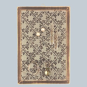 Puede incluir: Un organizador de joyas de madera marr&oacute;n con un fondo de malla beige. El organizador tiene un patr&oacute;n floral y est&aacute; lleno de pendientes, un anillo y un colgante.