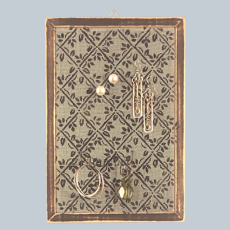 Puede incluir: Un organizador de joyas de madera marr&oacute;n con un fondo de malla beige. El organizador tiene un patr&oacute;n floral y est&aacute; lleno de pendientes, un anillo y un colgante.