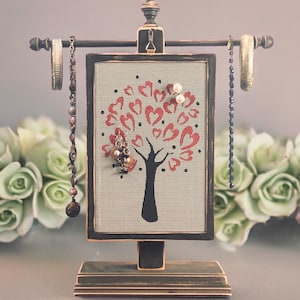 May include: A black wooden jewelry stand with a tree design featuring red hearts. The stand has a metal bar for hanging bracelets and a mesh panel for holding earrings.