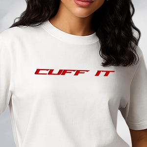 Peut inclure: Un t-shirt blanc avec le texte rouge "CUFF IT" sur la poitrine. Le t-shirt a des manches courtes et est associé à un jean noir. L'arrière-plan présente un ciel nuageux et une sculpture argentée réfléchissante.