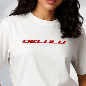 Peut inclure: Un t-shirt blanc avec le texte rouge "DELLULU" sur la poitrine. Le modèle porte un jean noir. L'arrière-plan présente un ciel nuageux et une sculpture argentée réfléchissante.